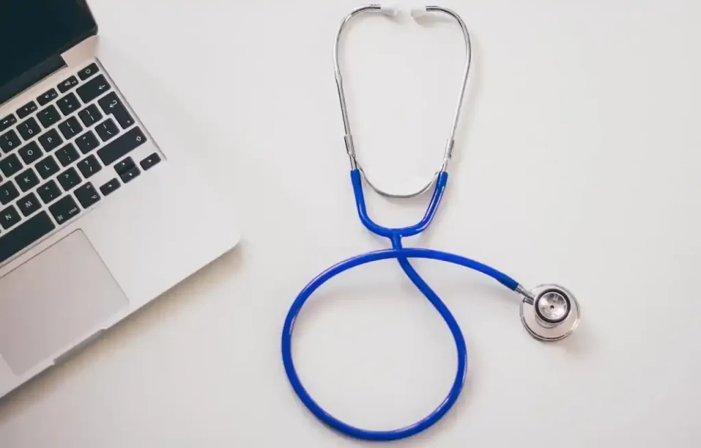 Estetoscópio azul ao lado de um notebook sobre mesa branca, representando consultas médicas e exame periódico ocupacional.