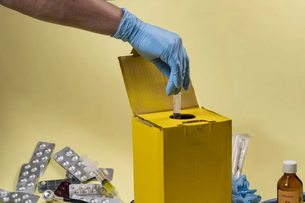 Mão com luva descartando material perfurocortante em caixa de descarte, representando riscos ocupacionais e segurança no trabalho.