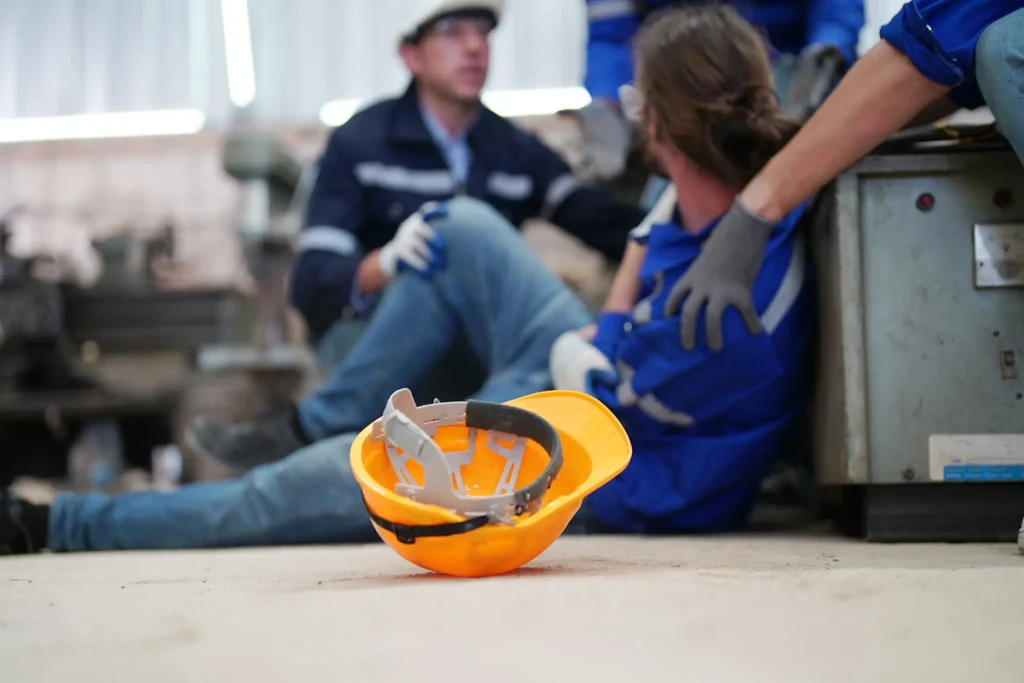 Trabalhador recebendo ajuda após acidente em fábrica enquanto colegas prestam apoio no local de trabalho